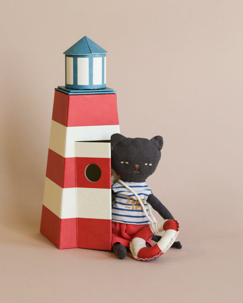 A handmade toy lighthouse with red and white stripes stands on a beige background. A Maileg Cat in Tower dressed as a lifeguard in a striped shirt and red shorts sits next to it, holding a small lifebuoy.