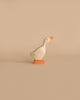 A minimalistic wooden duck figurine stands on a light beige surface against a matching beige background. This Holztiger Goose, Small is painted white with an orange beak and orange feet. Its simple design features a few subtle blue lines on its wings, embodying the charm of handcrafted wood, Made in Europe.