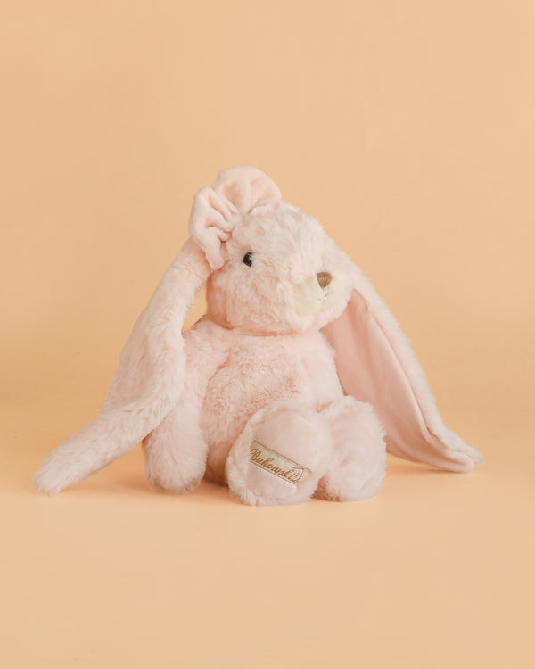 A soft, light pink Bukowski - Friendly Kanina - Misty Rose plush rabbit with long floppy ears sits against a matching peach background. This comforting companion features a stitched nose and a gold logo on its paw.