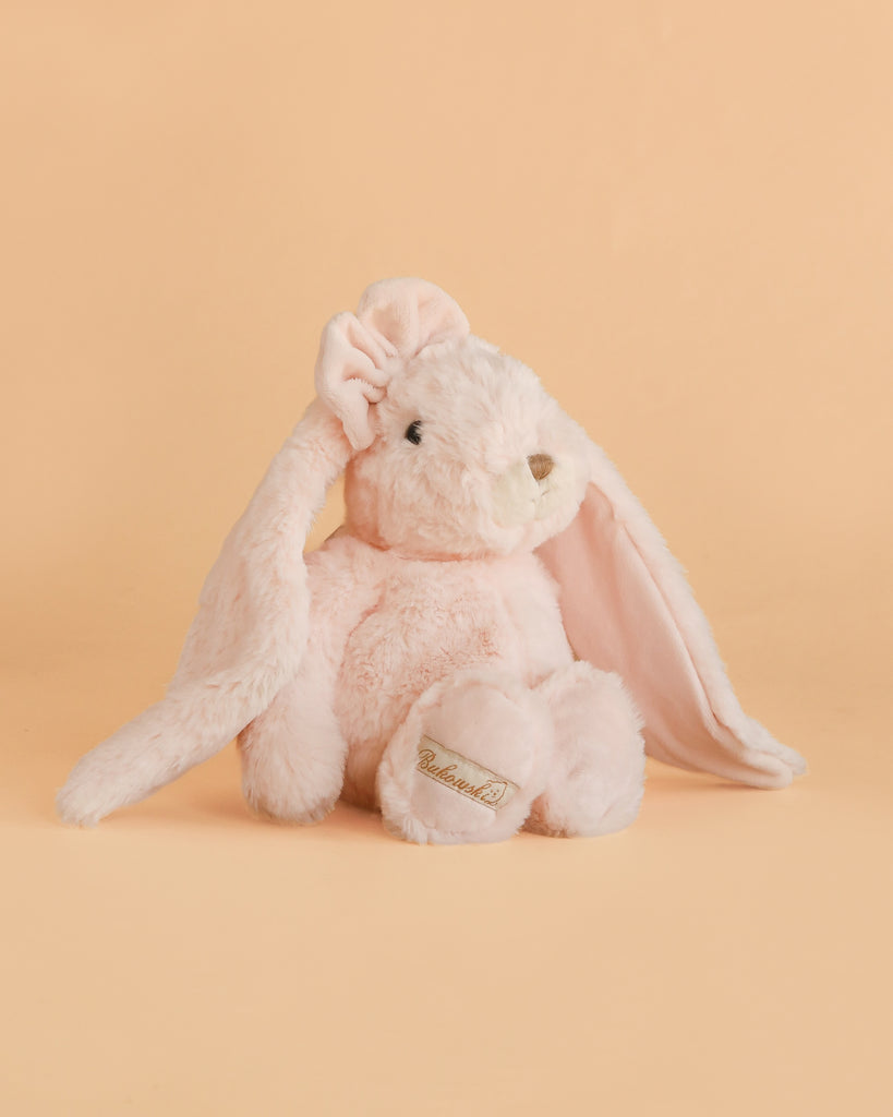 A soft, light pink Bukowski - Friendly Kanina - Misty Rose plush rabbit with long floppy ears sits against a matching peach background. This comforting companion features a stitched nose and a gold logo on its paw.