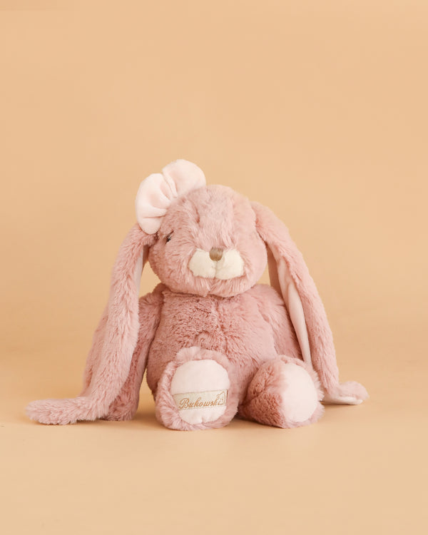 The Bukowski - Friendly Kanina - Antique Pink is a plush pink bunny with floppy ears, a soft pink bow on one ear, and “Beloved” embroidered on one paw. Sitting against a plain beige background, this comforting stuffed animal adds warmth and cheer to any space.