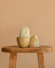 A Maileg Easter Egg With Chick, featuring a cream yellow plush chicken with a lace collar, sits inside an open decorative Easter egg on a wooden stool against a plain beige background.