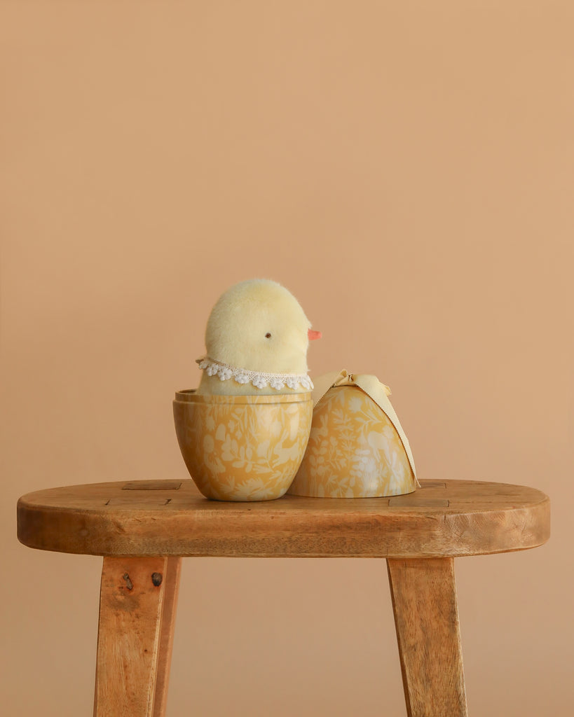 A Maileg Easter Egg With Chick, featuring a cream yellow plush chicken with a lace collar, sits inside an open decorative Easter egg on a wooden stool against a plain beige background.