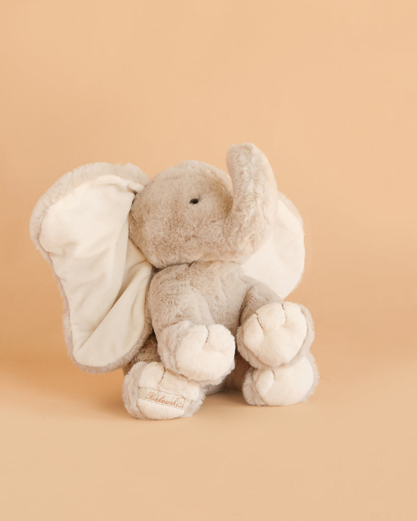 The Bukowski - August Elephant is a soft, light gray plush toy sitting upright on a beige background, featuring large floppy ears, a curved trunk, and embroidered detailing on its feet.