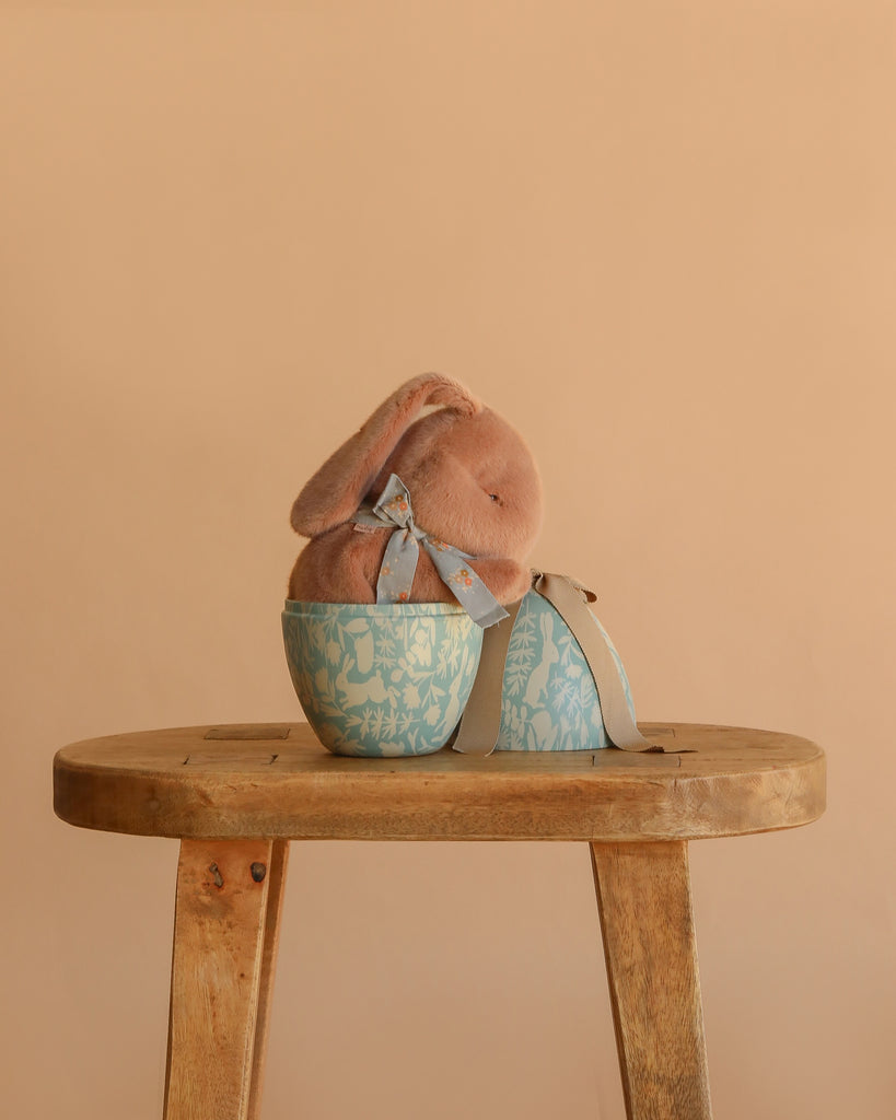 A Maileg Easter Egg With Sleeping Bunny featuring a soft brown plush bunny with a neck ribbon is displayed inside a decorative blue and white egg on a wooden stool, set against a plain beige background.