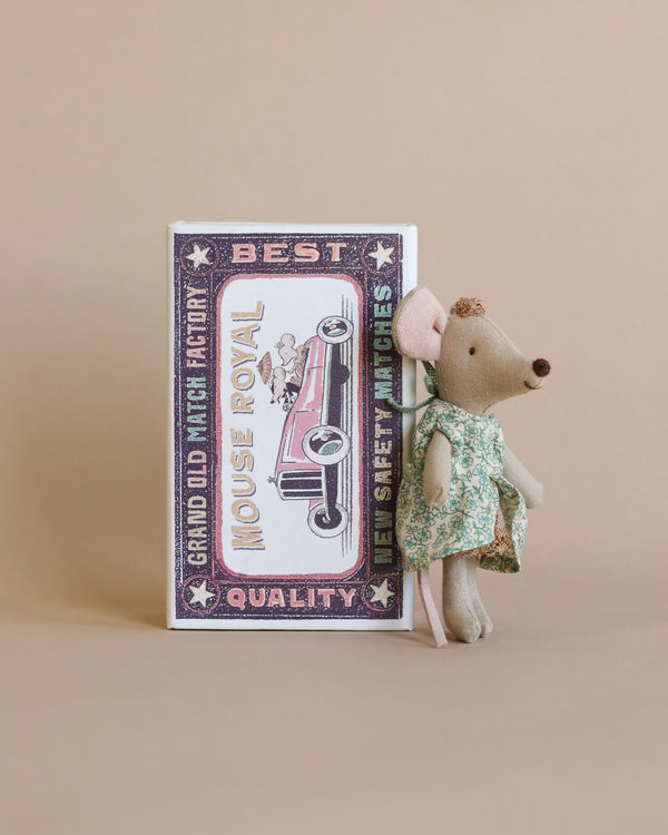 A Maileg Princess Little Sister mouse toy dressed in a green floral-patterned dress stands next to a vintage-style book with "mouse royal" and other text on it, set against a soft beige background.