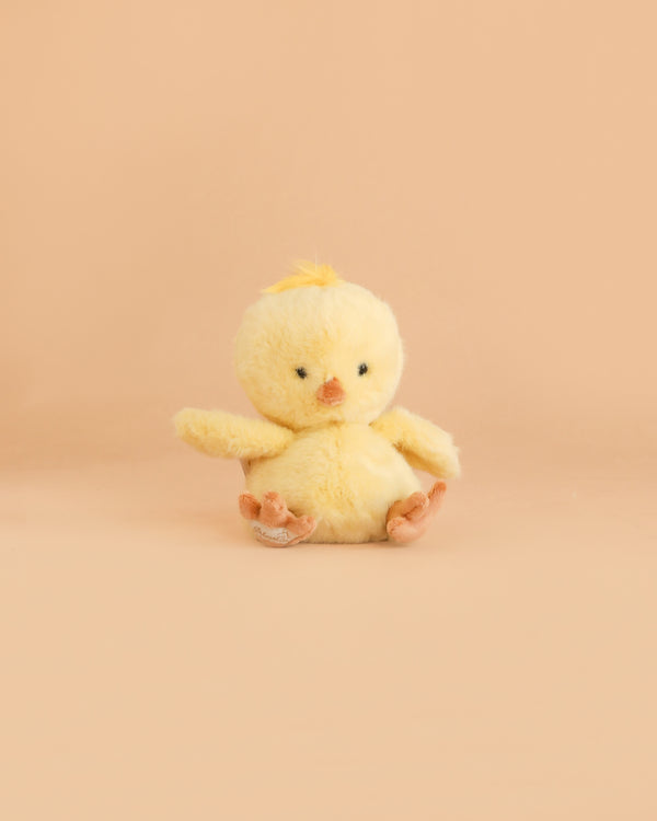 A Bukowski - Baby Chicky plush toy, small and yellow with black eyes, an orange beak and feet, sits facing forward on a light peach background.