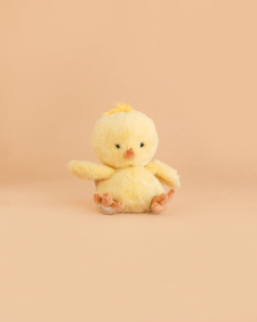 A Bukowski - Baby Chicky plush toy, small and yellow with black eyes, an orange beak and feet, sits facing forward on a light peach background.