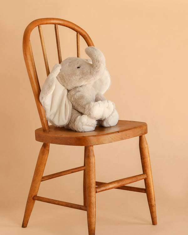 Bukowski - August Elephant, featuring soft gray fur, white ears, and feet, sits on a wooden chair against a light beige background—a perfect plush toy for bedtime cuddles.