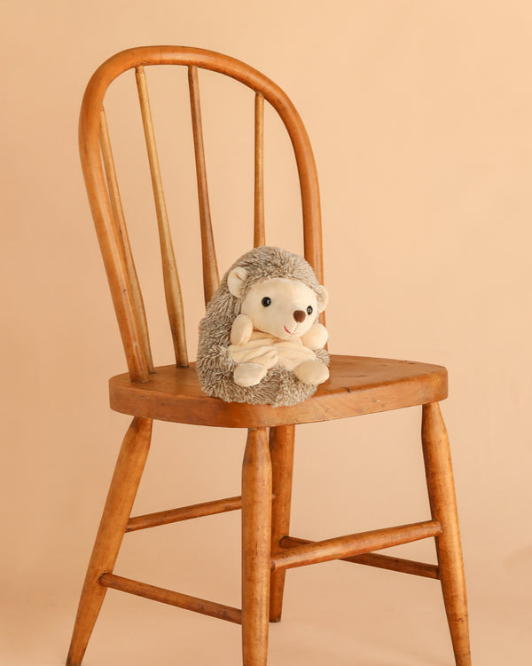 Bukowski - Sweet Hubert Hedgehog - Sitting is a safe stuffed animal, shown sitting on a wooden chair against a plain beige background—perfect as a cozy nap time toy.