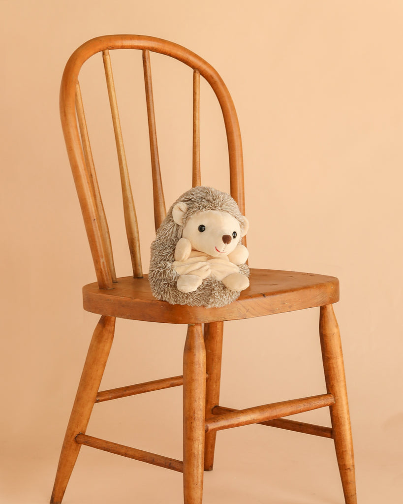 Bukowski - Sweet Hubert Hedgehog - Sitting is a safe stuffed animal, shown sitting on a wooden chair against a plain beige background—perfect as a cozy nap time toy.