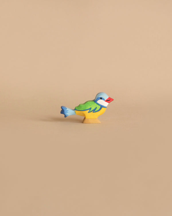 A small wooden bird figure painted in bright pastel colors stands against a plain beige background. The bird has a blue head and tail, green wings, yellow body, and a red beak. This delightful piece is reminiscent of the Holztiger Eurasian Blue Bird, reflecting the charm of handcrafted wood toys as it faces left.