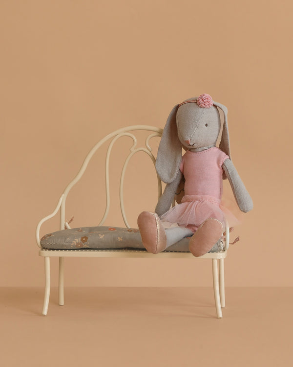 A Maileg Bunny, Size 2 - Classic, wearing a ballet suit and skirt in rose, sits on a small vintage-style cream bench with a floral cushion against a beige background.