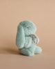 A soft, light blue Maileg Mini Plush Bunny - Mint sits on a beige background, facing away and looking over its shoulder, with a whimsical expression and a fluffy texture.