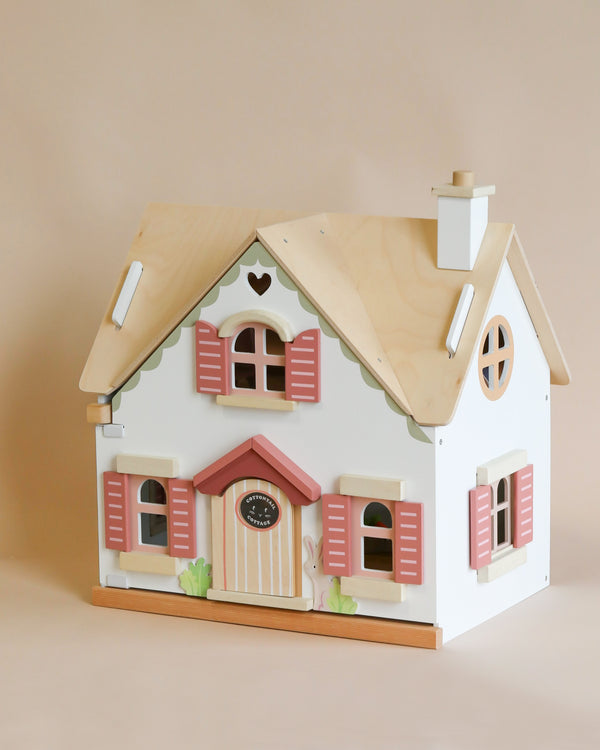 A Cottontail Cottage Dollhouse with a white exterior and pink shutters, featuring detailed windows, doors, and a natural wood roof, set against a pale beige background.
