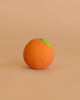 The Orange Baby Ball, shaped like a ripe orange with a green leaf, rests on a smooth light brown surface—its design evoking the gentle simplicity of sensory play against the matching background.