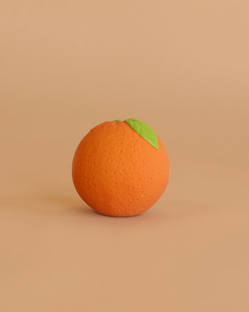 The Orange Baby Ball, shaped like a ripe orange with a green leaf, rests on a smooth light brown surface—its design evoking the gentle simplicity of sensory play against the matching background.
