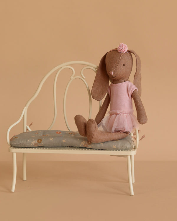 A Maileg Bunny, Size 1 in chocolate brown, dressed in a rose ballet suit and skirt, sits gracefully on a cream linen bench against a beige background.