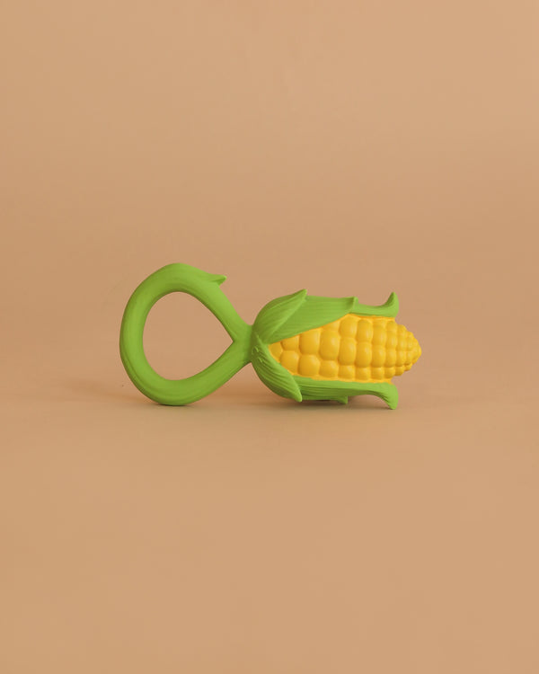 A Corn Rattle Toy Rattle, shaped like yellow corn with a green handle and green leaf accents, is placed on a plain beige background.