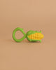 A Corn Rattle Toy Rattle, shaped like yellow corn with a green handle and green leaf accents, is placed on a plain beige background.