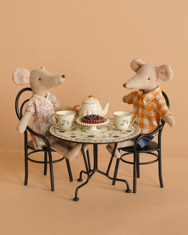 The Maileg Mom & Dad Afternoon Tea Set features plush mice seated at a bistro table, enjoying tea with pastries. One mouse wears a dress, the other a plaid shirt; black chairs set against a beige background complete the scene.