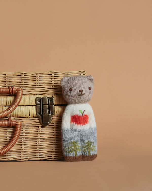 The Blabla Organic Alpaca Wool - Happy Go Lucky Bear Doll from Peru sports a white sweater with a red apple, blue pants, and stands next to a wicker basket with a latch on a beige background. Made from organic alpaca fleece for extra softness and charm.