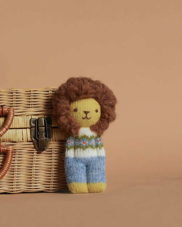 The Blabla Organic Alpaca Wool Happy Go Lucky Lion Doll is a handmade Peruvian plush toy with a fluffy brown mane, patterned sweater, and blue pants, crafted from organic alpaca fleece for softness. Shown by a wicker basket on a beige background.