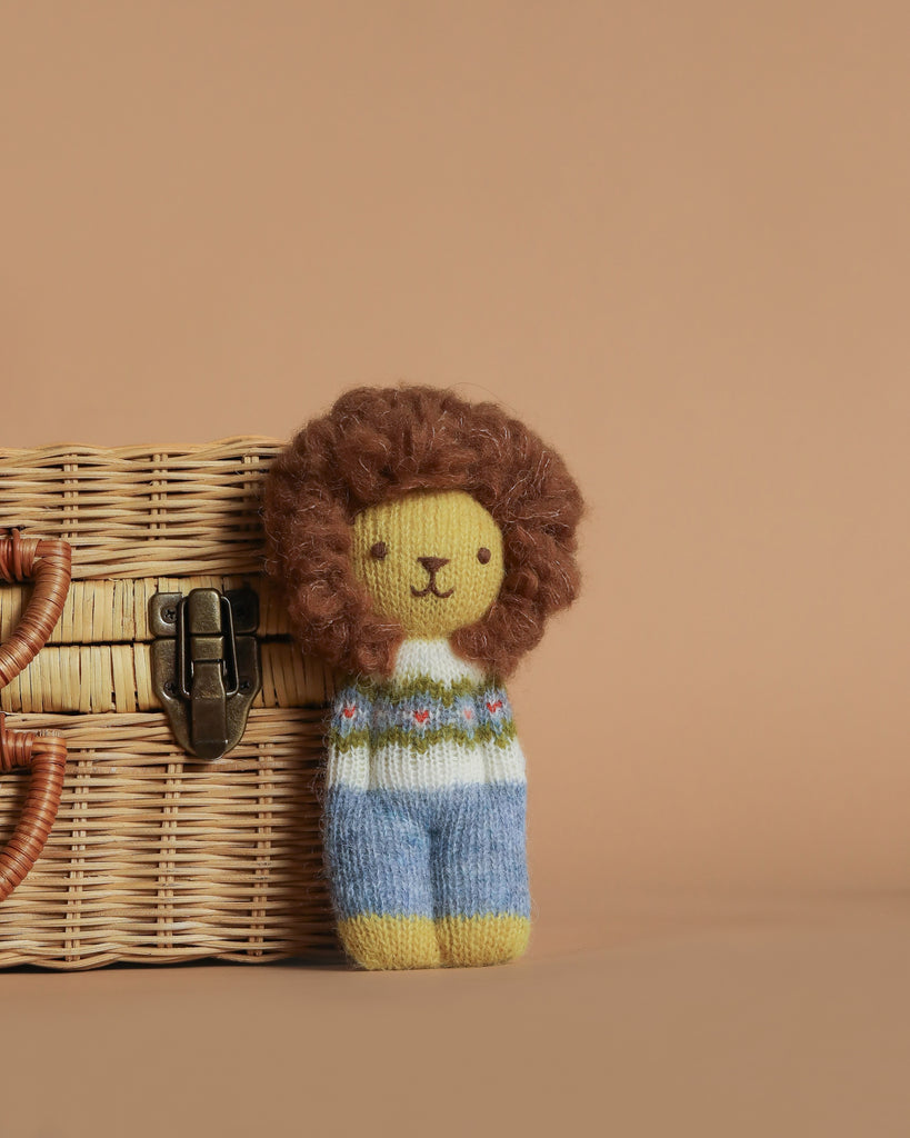 The Blabla Organic Alpaca Wool Happy Go Lucky Lion Doll is a handmade Peruvian plush toy with a fluffy brown mane, patterned sweater, and blue pants, crafted from organic alpaca fleece for softness. Shown by a wicker basket on a beige background.