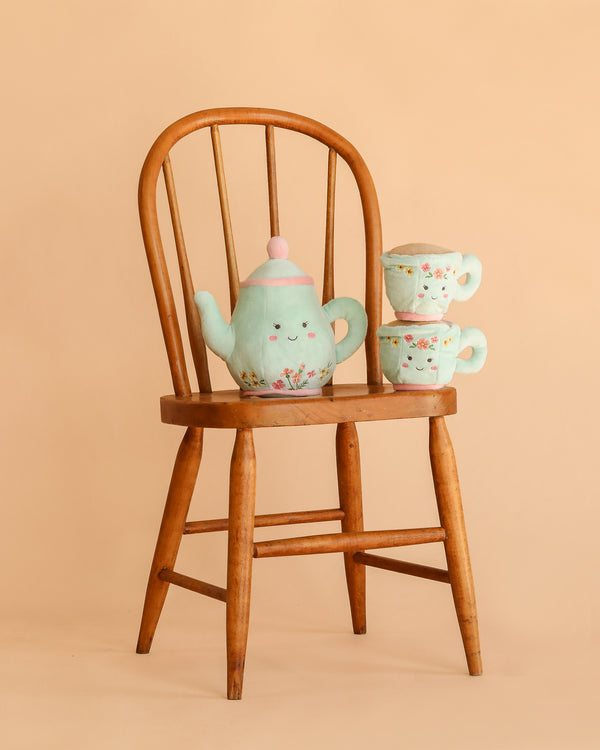 The Mon Ami - Afternoon Tea Set Plush Toy is displayed on a wooden chair, featuring a charming pastel teapot with a happy face and floral details, alongside two stacked matching teacups with cheerful smiles, all against a beige background—perfect for creating an adorable pretend play tea set or cuddly tea party toys display.