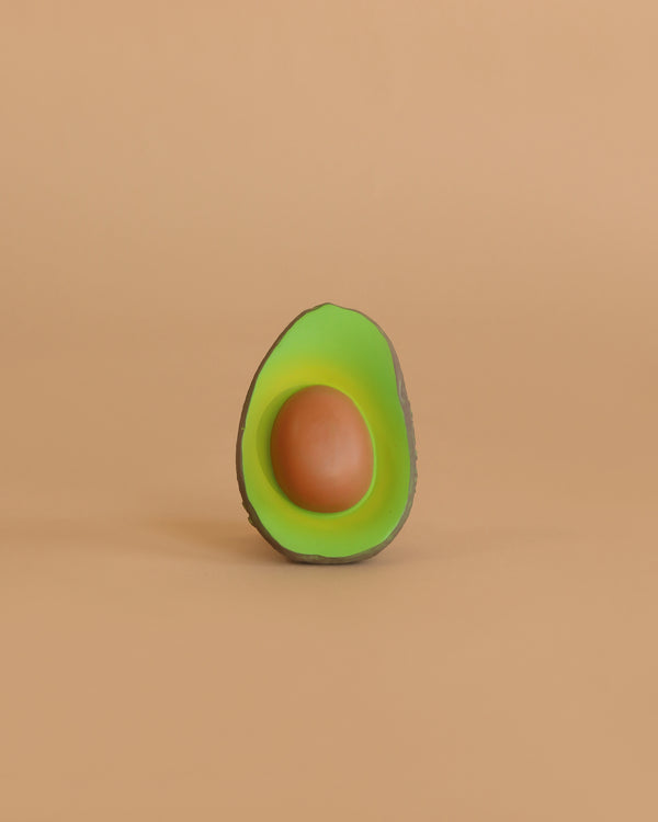 Arnold the Avocado Teething Toy, shaped like a halved avocado with green flesh and a brown seed, sits centered on a solid beige background—its natural rubber design makes it perfect for sensory play.