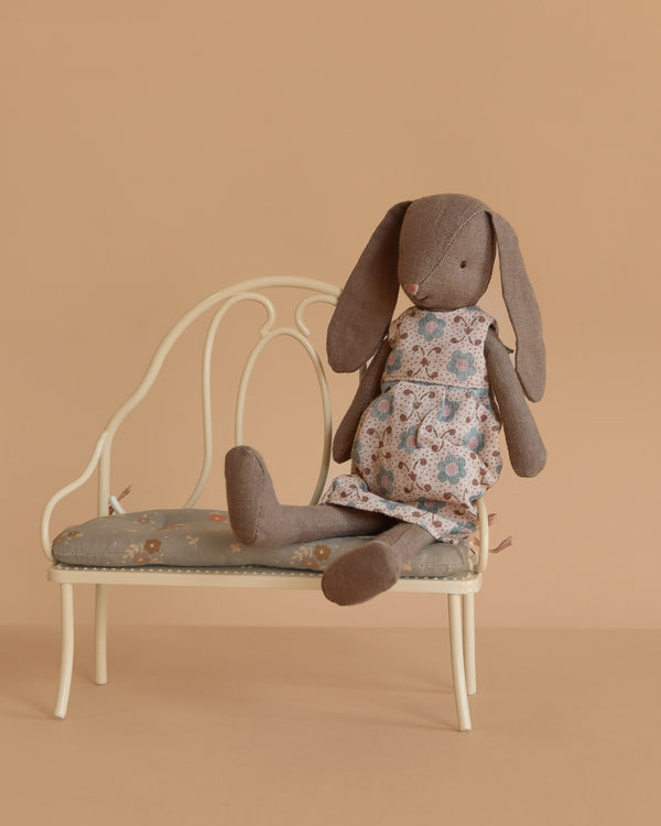The Maileg Bunny, Size 2 - Brown - Dress, sits on a vintage-style cream bench with a padded seat against a plain beige background.