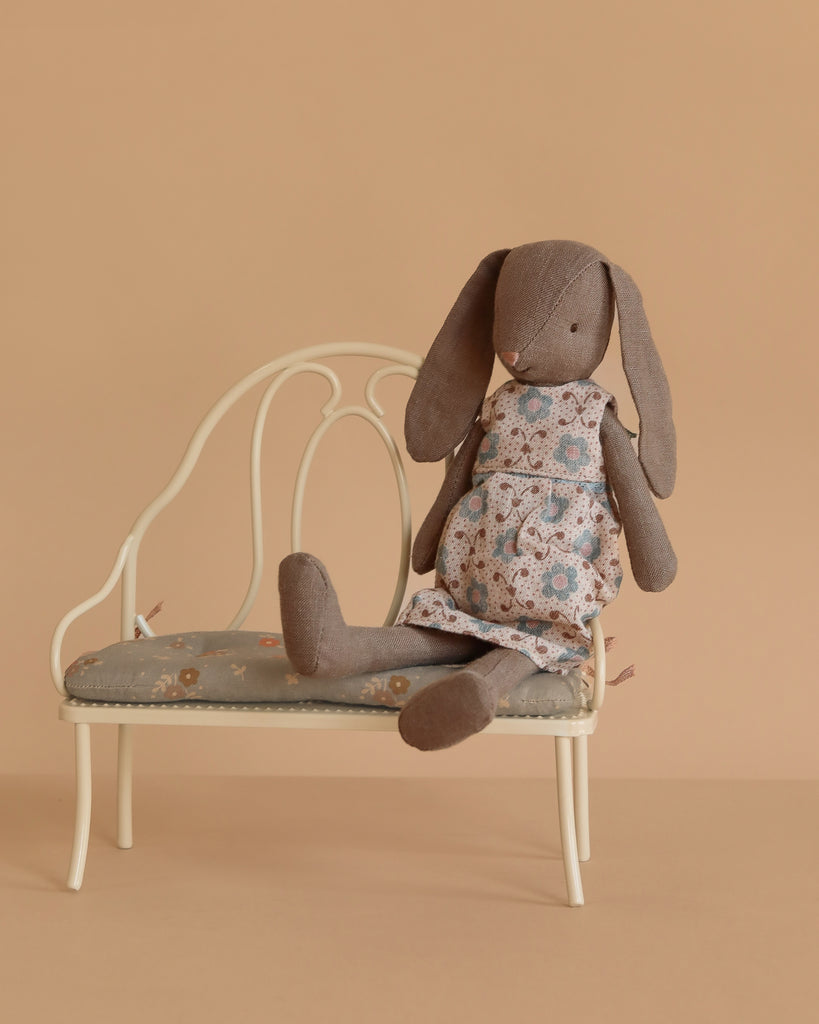 The Maileg Bunny, Size 2 - Brown - Dress, sits on a vintage-style cream bench with a padded seat against a plain beige background.