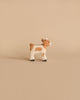 A small, handcrafted wooden figurine of a calf, painted white with light brown patches, black hooves, and a short tail, stands on a solid beige background. Made in Europe by Holztiger Baby Goat.