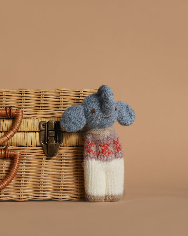 The Blabla Organic Alpaca Wool - Happy Go Lucky Elephant Doll is a handmade Peruvian toy with blue ears and trunk, a brown and white body with red patterns, crafted from organic alpaca fleece, shown by a wicker basket on a plain brown background.