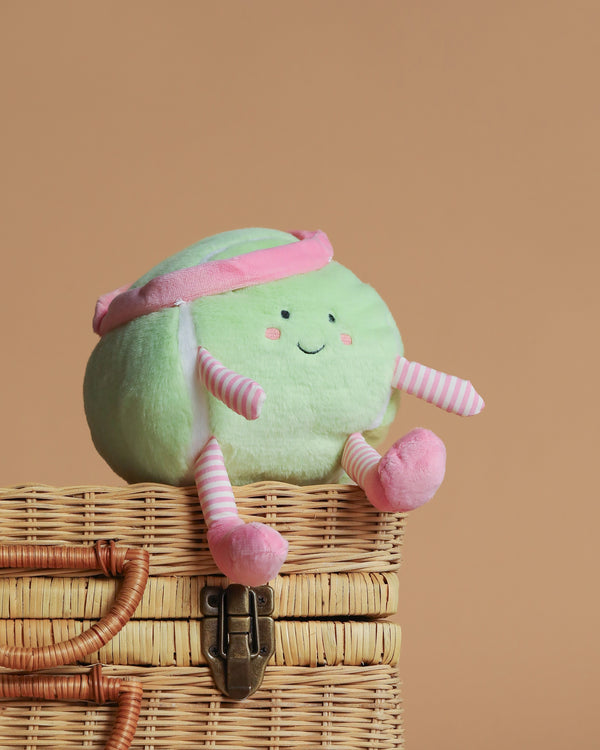 The Mon Ami - Tina Tennis Ball Plush Toy, with a smiling face, pink headband, and pink-striped arms and legs, sits atop a closed wicker basket against a plain beige background.