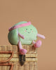 The Mon Ami - Tina Tennis Ball Plush Toy, with a smiling face, pink headband, and pink-striped arms and legs, sits atop a closed wicker basket against a plain beige background.