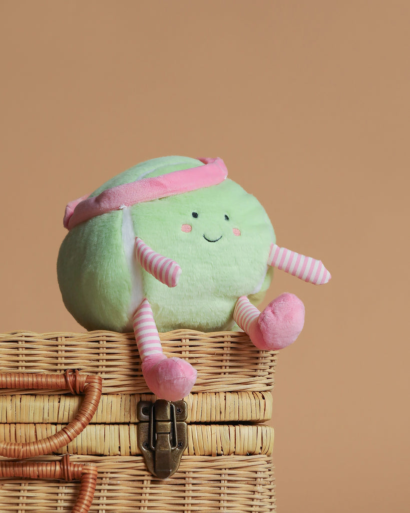 The Mon Ami - Tina Tennis Ball Plush Toy, with a smiling face, pink headband, and pink-striped arms and legs, sits atop a closed wicker basket against a plain beige background.