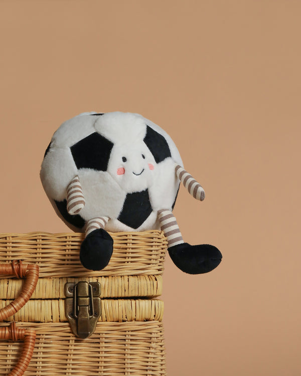 The Mon Ami - Sergio Soccer Ball Plush Toy, featuring a smiling face, striped arms and legs, and black feet, sits on a woven basket against a beige background—a cute soccer gift for any fan.