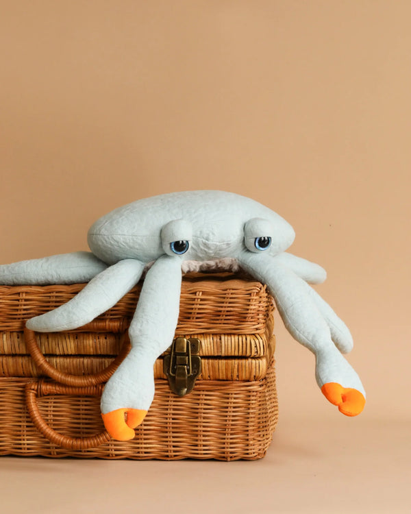 A large, light blue BigStuffed Mini Crab - Blue resembling a crab with orange tips on its claws is placed on top of a closed, wicker basket against a beige background. Handcrafted with a soft, plush texture and a relaxed expression, this adorable crab is safe for newborns.