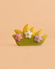 A small decorative piece called the Bumbu Handmade Wooden Blossom Bush resembles a charming wooden toy, featuring green and yellow grass shapes and three pastel flowers—pink, white, and purple—on a peach background.