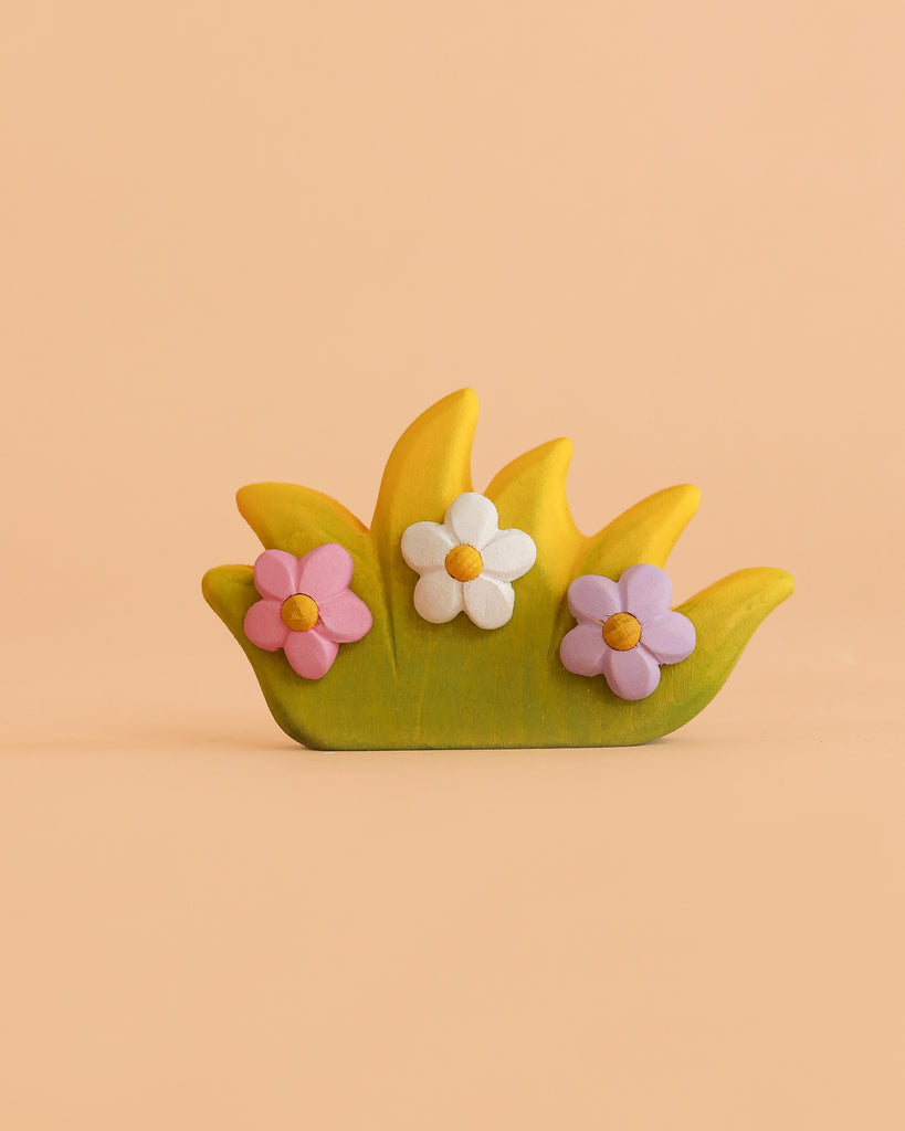 A small decorative piece called the Bumbu Handmade Wooden Blossom Bush resembles a charming wooden toy, featuring green and yellow grass shapes and three pastel flowers—pink, white, and purple—on a peach background.
