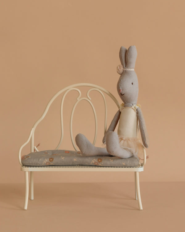 A Maileg Rabbit, Size 1 - Classic, dressed in a cream ballet suit and skirt, sits on a small cotton linen bench with a floral cushion against a solid beige background and floor.