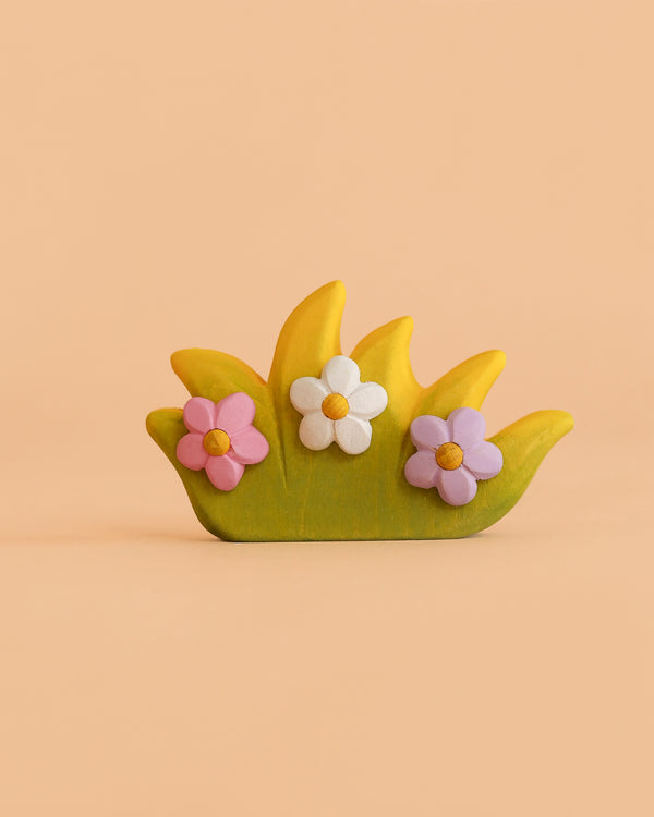 The Bumbu Handmade Wooden Blossom Bush is a small, decorative object shaped like green grass with yellow tips and adorned with three pastel flowers in pink, white, and purple. Set against a solid peach background, this Montessori-inspired wooden toy brings a charming and educational accent to any space.
