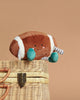 The Mon Ami Freddie Football Plush Toy, shaped like an American football with a smiling face, teal arms, and striped legs, sits on a woven basket against a beige background—a cute cuddle buddy and perfect gift for kids.