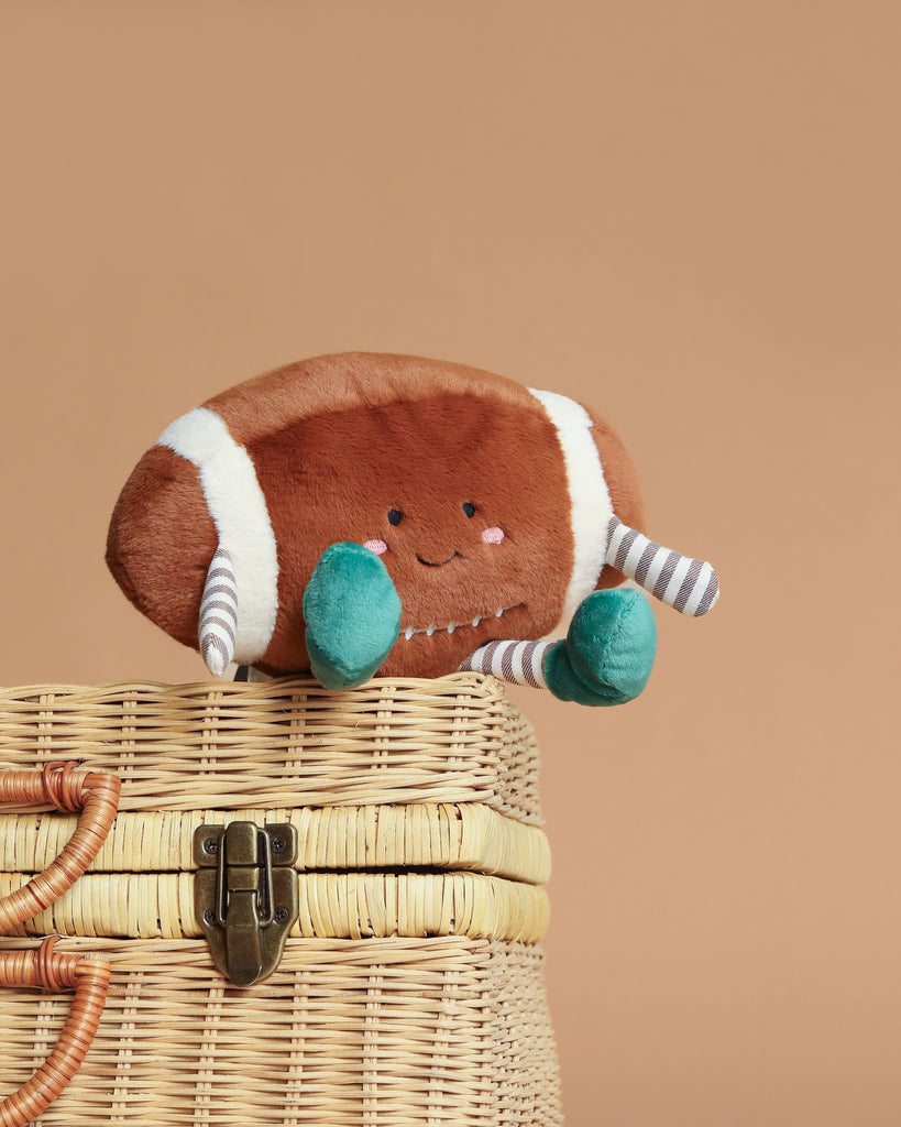 The Mon Ami Freddie Football Plush Toy, shaped like an American football with a smiling face, teal arms, and striped legs, sits on a woven basket against a beige background—a cute cuddle buddy and perfect gift for kids.