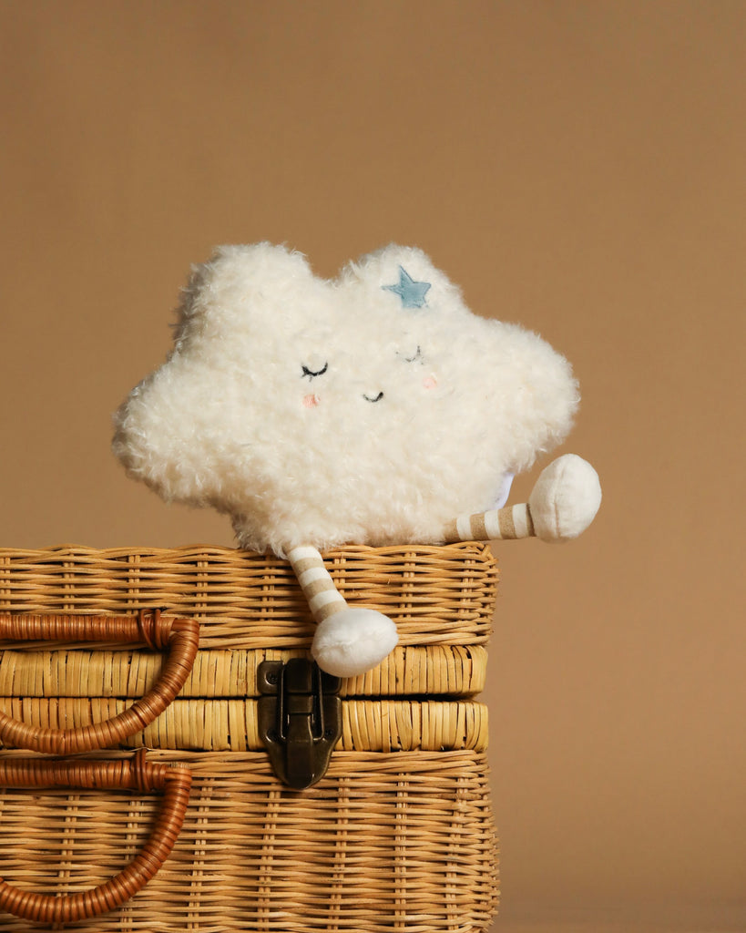 The Mon Ami - Soft Cloud Plush Toy, featuring closed eyes, a blue star, and striped legs, sits on a wicker basket against a brown background—an irresistibly cozy accent for any room.