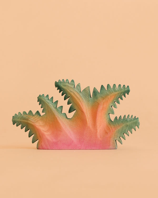 The Bumbu Handmade Wooden Fern Bush—a handcrafted toy painted in pink, orange, and green—features spiky, abstract foliage and rests against a plain peach background, making it a unique piece of decor as well as an educational plant toy for curious minds.
