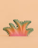 The Bumbu Handmade Wooden Fern Bush—a handcrafted toy painted in pink, orange, and green—features spiky, abstract foliage and rests against a plain peach background, making it a unique piece of decor as well as an educational plant toy for curious minds.