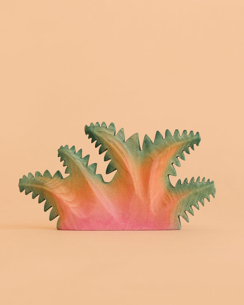 The Bumbu Handmade Wooden Fern Bush—a handcrafted toy painted in pink, orange, and green—features spiky, abstract foliage and rests against a plain peach background, making it a unique piece of decor as well as an educational plant toy for curious minds.