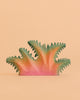 The Bumbu Handmade Wooden Fern Bush is a handcrafted wooden toy, painted in pink and green, with abstract spiky shapes reminiscent of fern foliage, pictured against a plain peach background.
